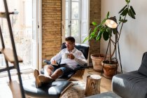 Hombre de negocios serio sentado en sillón en casa y escribiendo planes en organizador mientras disfruta del café durante el descanso - foto de stock