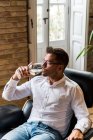 Pensive male resting in comfortable leather armchair with glass of white wine and looking away in thoughts — Stock Photo
