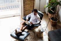 Зверху чоловічий підприємець сидить у кріслі і працює в Інтернеті над проектом, використовуючи мобільний телефон і друкуючи на ноутбуці — стокове фото
