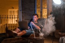 Расслабленный мужчина сидит в удобном кресле с ноутбуком и курит кальян во время просмотра фильма и наслаждаясь выходными — стоковое фото