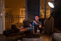 Entspannter Mann sitzt im bequemen Sessel mit Laptop und raucht Wasserpfeife, während er Film schaut und das Wochenende genießt — Stockfoto