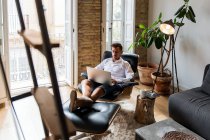 Красивый мужчина-предприниматель сидит дома в кресле и работает над проектом на ноутбуке, наслаждаясь кофе — стоковое фото