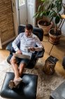 Красивий чоловік-підприємець сидить у кріслі вдома і працює над проектом на ноутбуці, насолоджуючись кавою — стокове фото