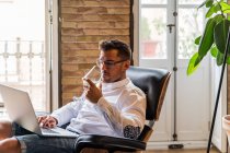 Beau entrepreneur masculin assis dans un fauteuil à la maison et travaillant sur un projet sur ordinateur portable tout en appréciant le vin — Photo de stock