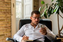 Geschäftsmann sitzt im Sessel und macht sich Notizen, während er im Smartphone surft und Nachrichten checkt — Stockfoto