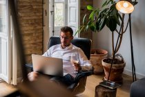 Schöner männlicher Unternehmer sitzt zu Hause im Sessel und arbeitet am Laptop an einem Projekt, während er Wein genießt — Stockfoto