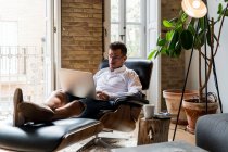 Bonito empresário masculino sentado em poltrona em casa e trabalhando no projeto no laptop enquanto desfruta de café — Fotografia de Stock