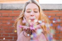 Happy young female with long blond hair blowing colorful confetti from hands while standing near brick wall on sunny day — Stock Photo