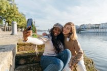 Zufriedene beste Freundinnen sitzen auf der Treppe an der Promenade und machen bei einem Spaziergang an einem sonnigen Tag in der Stadt ein Selfie mit dem Smartphone — Stockfoto