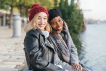 Vista lateral de alegres amigas multiétnicas no calçadão enquanto desfrutam de fim de semana juntas no outono e olhando para a câmera — Fotografia de Stock