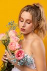 Молода неемоційна красива жінка в білій сукні з голими плечима, що тримає ніжні рожеві троянди, стоячи з закритими очима на жовтому тлі в фотостудії — стокове фото
