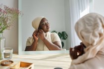 Rückansicht einer selbstbewussten jungen Afrikanerin mit traditionellem Kopftuch, die in den Spiegel schaut und im modernen Schlafzimmer Ohrringe anlegt — Stockfoto