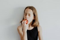Girl in casual top closed eyes while biting fresh ripe red apple against white background — Stock Photo
