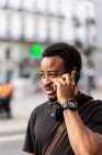 Jovem conteúdo afro-americano masculino em relógio de pulso falando no celular enquanto olha para longe na cidade — Fotografia de Stock