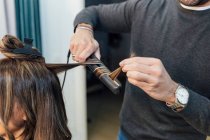 Crop estilista masculino irreconhecível em roupas casuais usando alisador de cabelo ao fazer cachos para o cliente feminino no salão de beleza — Fotografia de Stock