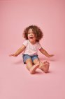 Cute cheerful toddler girl with curly hair in casual clothes having fun looking away smiling while sitting on pink background — Stock Photo