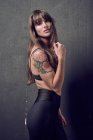 Sensual female with tattoo wearing bra touching neck standing in studio against black background — Stock Photo