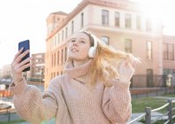 Впевнена молода жінка в теплому светрі і навушники, які торкаються довгохвильового волосся, приймаючи селфі на смартфон у сонячний день у місті — стокове фото