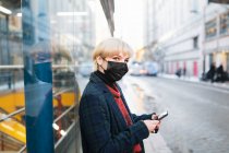 Вид сбоку спокойная молодая женщина в теплом пальто и защитной маске для лица просматривает современный мобильный телефон, стоя на улице города в зимний день в Мадриде, Испания — стоковое фото