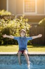 Kid sitting on edge of outdoor swimming pool and splashing water with legs on sunny summer day — Stock Photo