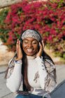 Восхитительная афроамериканка сидит в парке и слушает музыку в наушниках, наслаждаясь песнями с закрытыми глазами — стоковое фото