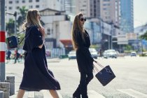 Vue latérale de jeunes partenaires féminines élégantes blondes dans des vêtements à la mode se promenant sur la route asphaltée en ville — Photo de stock