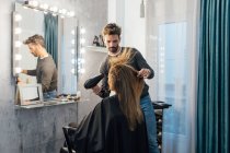 Этнический мужчина парикмахер сушит волосы клиентки с закрытыми глазами в современной студии красоты — стоковое фото