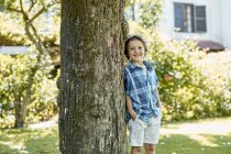 Feliz niño con estilo en pantalones cortos y camisa a cuadros apoyado en el tronco del árbol y sonriendo mientras descansa en el patio trasero en el día soleado - foto de stock