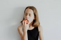 Chica en la parte superior casual ojos cerrados mientras muerde manzana roja madura fresca contra fondo blanco - foto de stock