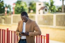 Concentrado jovem afro-americano masculino em roupa da moda inclinando-se sobre cerca na rua e navegação celular no dia ensolarado de verão — Fotografia de Stock