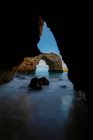 Through cave picturesque view of arched rocky formations in ocean coastline under blue sky in Praia da Abandeira, Algarve Portugal — Stock Photo