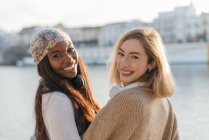 Vor der Kamera zwei multirassische Freundinnen mit Kopfhörern, die an einem sonnigen Tag gemeinsam auf einem Damm genießen — Stockfoto