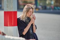 Вид збоку молодої жінки в шарфі куріння цигарки біля поста на міській дорозі в спині освітлений — стокове фото