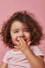 Cute cheerful toddler girl with curly hair in casual clothes covering mouth with hand while looking away smiling on pink background — Stock Photo