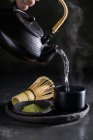 Обрізати анонімну людину, що поливає гарячу воду з чайника, готуючись до чайної церемонії з порошком мачі на тарілці поблизу шасена — стокове фото