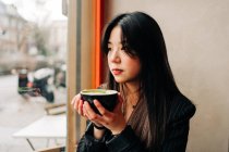 Morena de pelo largo Asiática tomando un café en una cafetería mientras mira un celular por la ventana - foto de stock