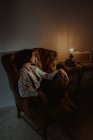 Vista lateral da mulher serena relaxando na poltrona de couro velho no quarto vintage com lâmpada brilhante e olhando para longe — Fotografia de Stock