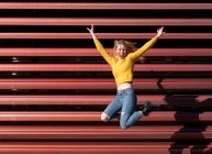 Full body of cheerful young female millennial in casual outfit jumping high with raised arms and smiling near metal fence on street — Stock Photo