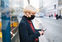 Вид сбоку спокойная молодая женщина в теплом пальто и защитной маске для лица просматривает современный мобильный телефон, стоя на улице города в зимний день в Мадриде, Испания — стоковое фото