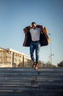 Cuerpo completo de hombre joven afroamericano en traje elegante ajustando el abrigo de moda mientras camina abajo en el parque de la ciudad contra el cielo azul sin nubes - foto de stock