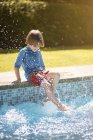 Неузнаваемый ребенок сидит на краю открытого бассейна и брызгает водой с ногами в солнечный летний день — стоковое фото