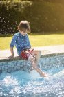 Niño irreconocible sentado en el borde de la piscina al aire libre y salpicaduras de agua con las piernas en el día de verano soleado - foto de stock