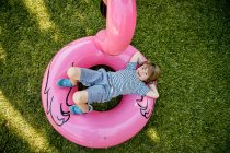 Повне тіло веселого маленького хлопчика в повсякденному одязі лежить на надувному рожевому фламінго, розважаючись на трав'янистому газоні в парку — стокове фото