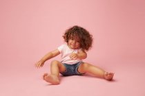 Mignon joyeux bambin fille avec des cheveux bouclés dans des vêtements décontractés avoir du plaisir à regarder loin en souriant tout en étant assis sur fond rose — Photo de stock