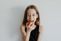 Cheerful preteen girl in casual top smiling while biting fresh ripe red apple against white background — Stock Photo