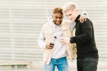 Heureux jeune Afro-Américain en vêtements à la mode souriant tout en se tenant dans la rue avec la main sur l'épaule d'un ami masculin montrant la vidéo sur smartphone — Photo de stock