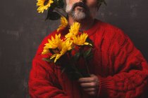 Hombre maduro creativo irreconocible en suéter de punto que cubre la cara con girasoles brillantes sobre fondo negro - foto de stock