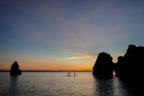 Atletas anónimos praticam desporto de prancha perto de silhuetas de formação rochosa em mar ondulado sob céu nublado no crepúsculo no Algarve Portugal — Fotografia de Stock