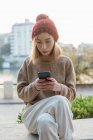Молода жінка в повсякденному вбранні, сидячи на кам'яному паркані та смс-повідомленні на мобільному телефоні, розважаючись у вихідні у місті — стокове фото