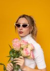 Joven hermosa hembra en traje blanco y gafas de sol de moda sosteniendo delicadas rosas rosadas mientras está de pie sobre fondo amarillo en el estudio de fotos - foto de stock
